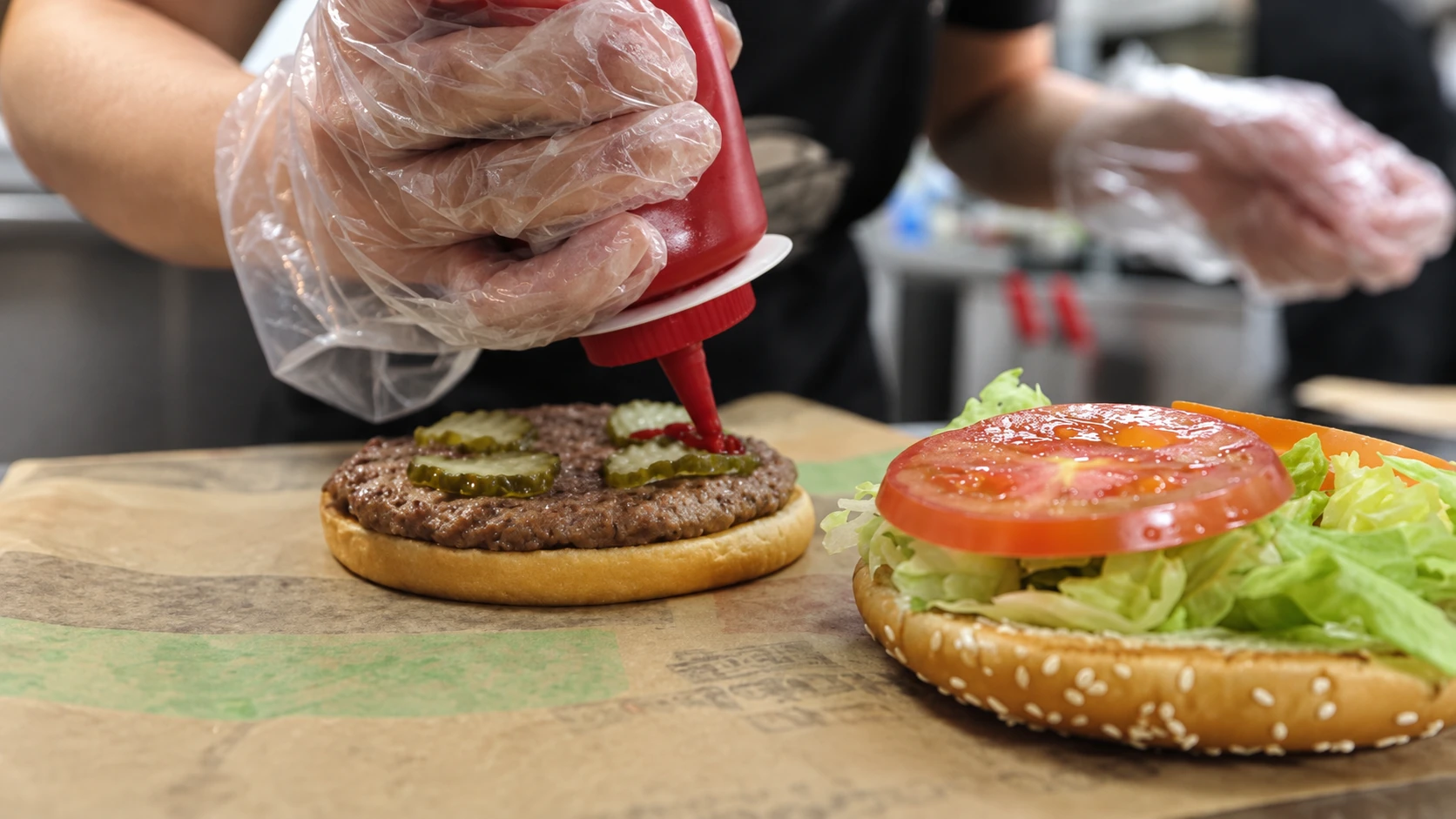 Gloved hands adding sauce to a burger beside lettuce and sliced tomato.