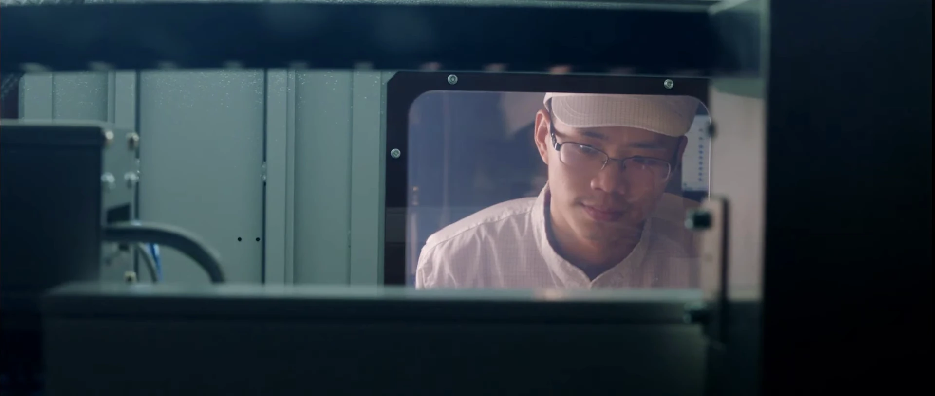 A technician in a cap and glasses viewed through manufacturing equipment.