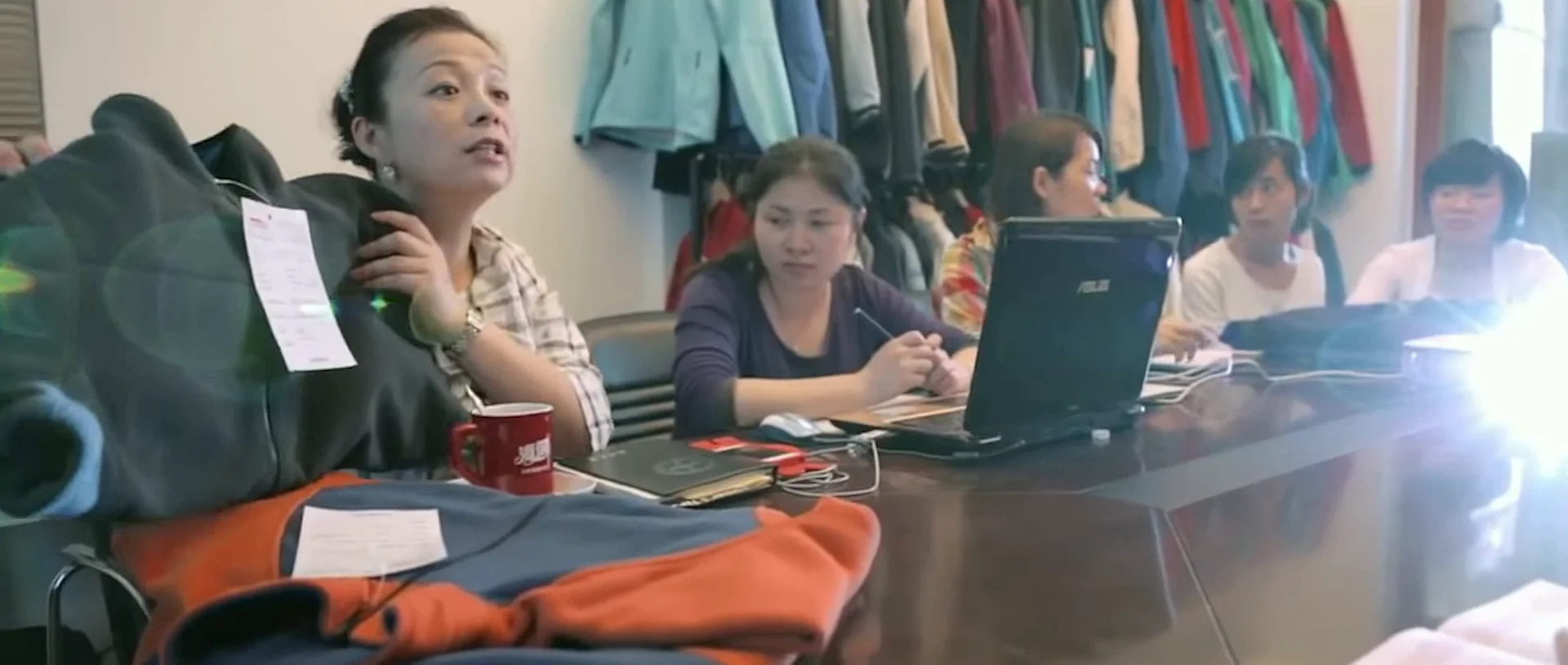 Women reviewing garments, paperwork and a laptop in a small workroom.