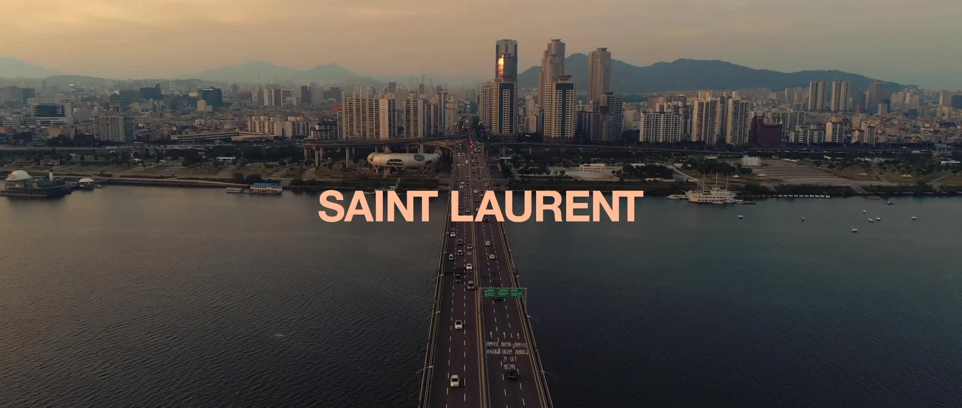 Saint Laurent title over a river, bridge and city skyline at dusk.
