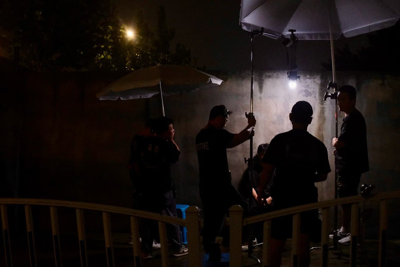 Several people stand around light stands and a hanging lamp beneath umbrellas at night.