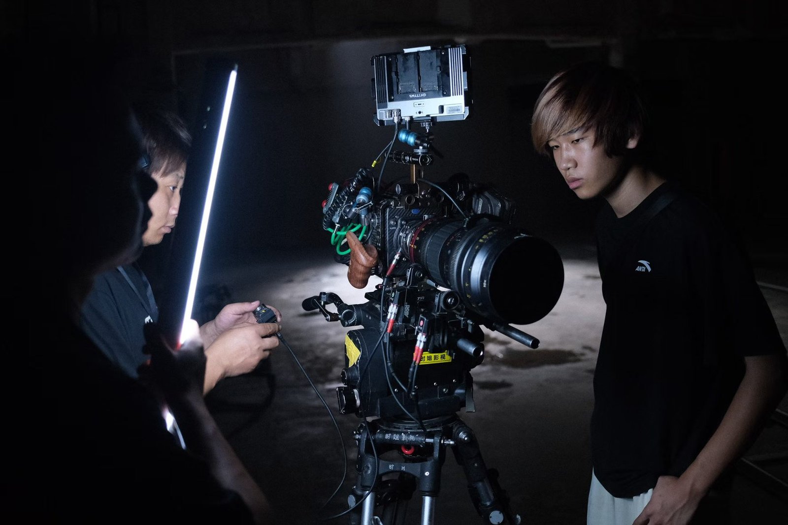Two people stand beside a cinema camera rig with a lens, cables and an external monitor.