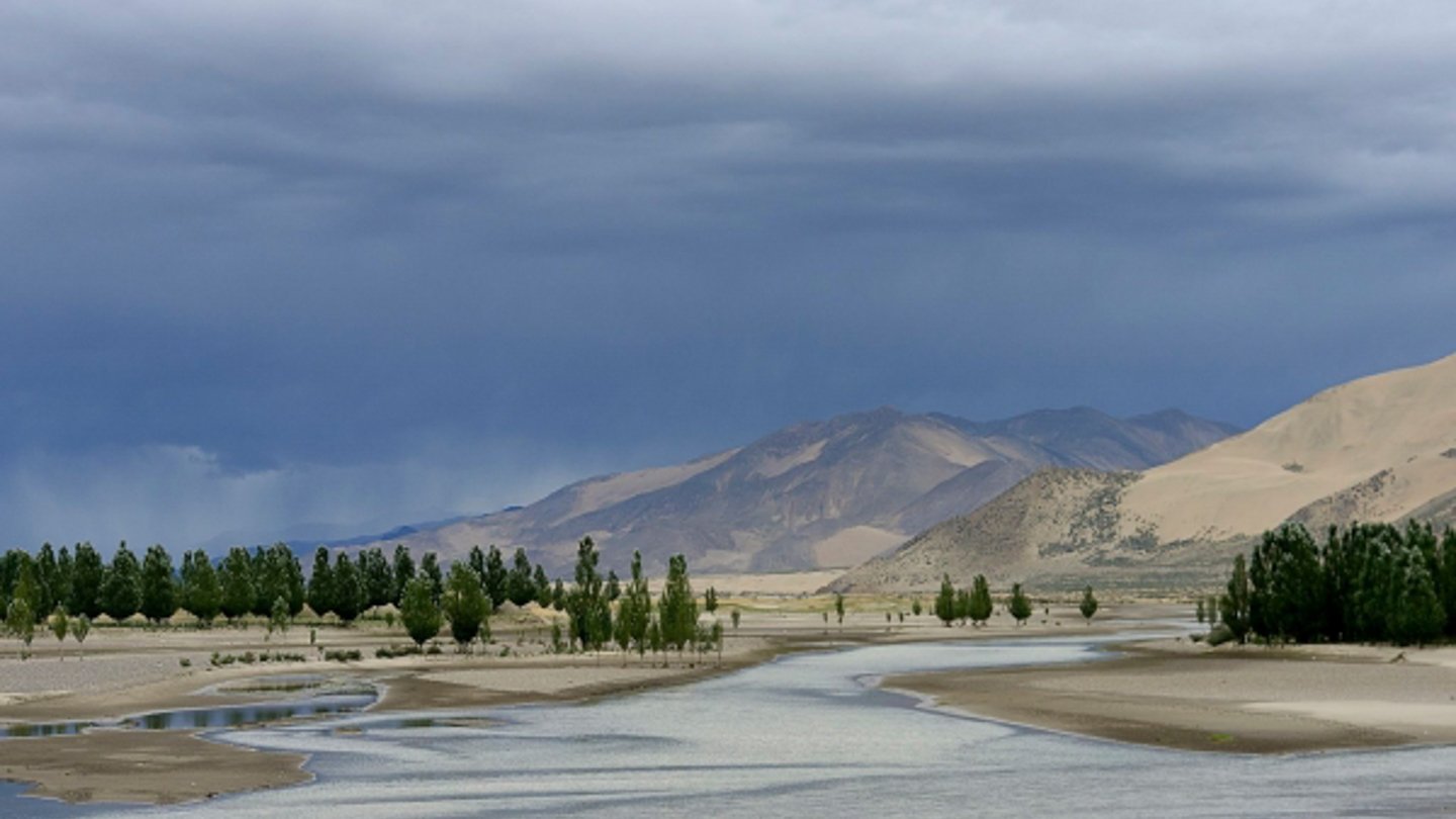 Winding water and tree-lined banks beneath a mountain sky. Programme still.