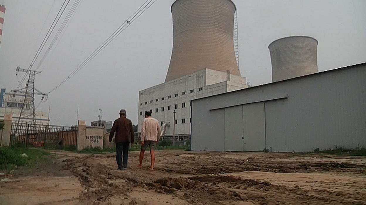 Two figures walking beside an industrial complex with cooling towers. Film still.