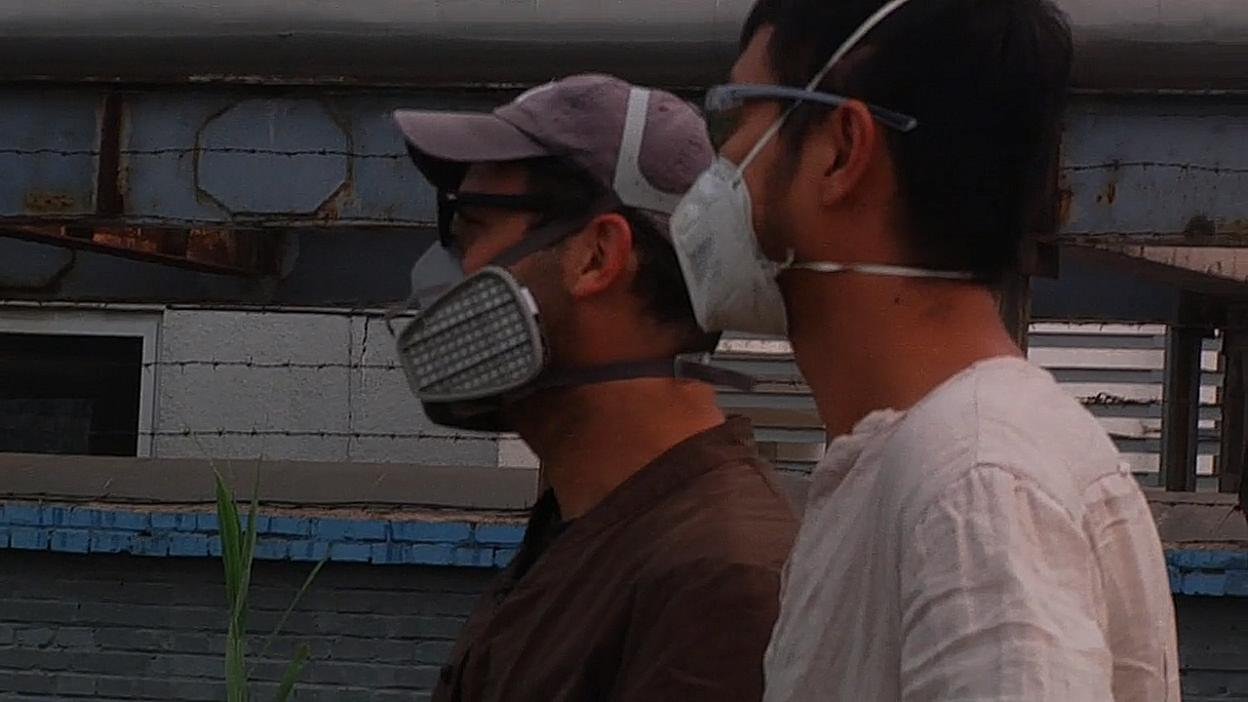 Two figures wearing respirators beside an industrial wall. Film still.