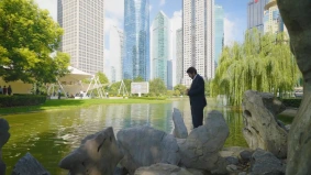 Man in a business suit standing by a serene lake in a modern city park with skyscrapers in the background.
