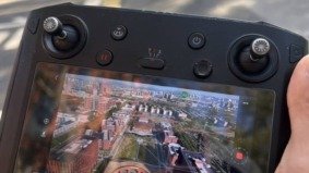 Hand holding a drone remote controller displaying a live aerial view of city buildings and roads.