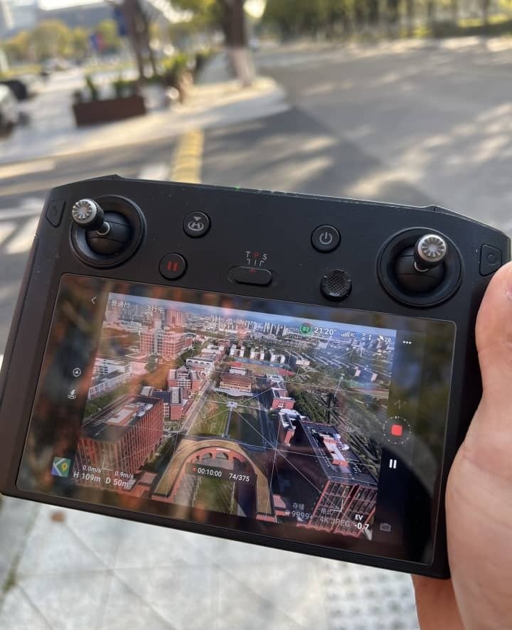 Hand holding a drone remote controller displaying a live aerial view of city buildings and roads.